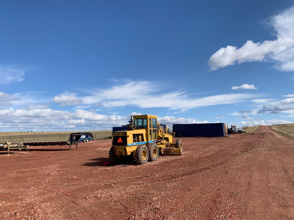 A yellow tractor is on the dirt in front of some buildings.